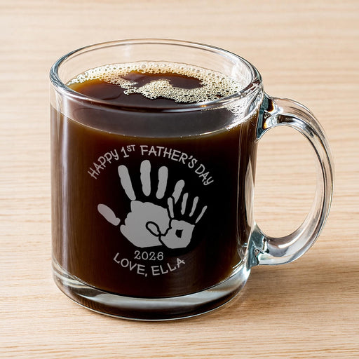 A glass mug filled with a dark liquid, likely coffee, with a handprint design engraved on it.