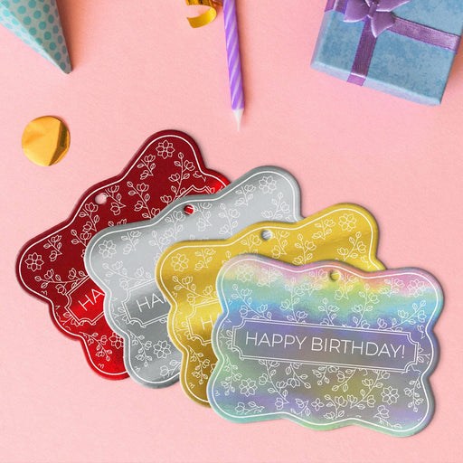 Four floral birthday gift tags in metallic gold, metallic silver, metallic red, and metallic rainbow lying beside colorful gifts and party decorations.