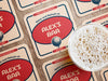 Custom square pulpboard coasters are shown laid out on a wooden surface. A beer mug is shown on top of a few of the coasters. The custom coasters feature a 19th hole bar design with an illustration of a golf ball. The tagline, bar name, established year, and location can be customized on these.