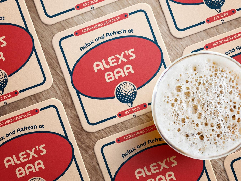 Custom square pulpboard coasters are shown laid out on a wooden surface. A beer mug is shown on top of a few of the coasters. The custom coasters feature a 19th hole bar design with an illustration of a golf ball. The tagline, bar name, established year, and location can be customized on these.