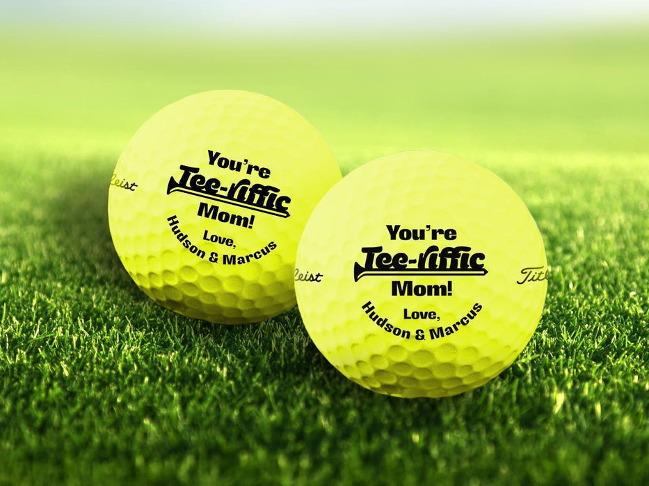 Two personalized yellow golf balls with a You're Tee-riffic Mom! design printed on them, placed on a grassy field.