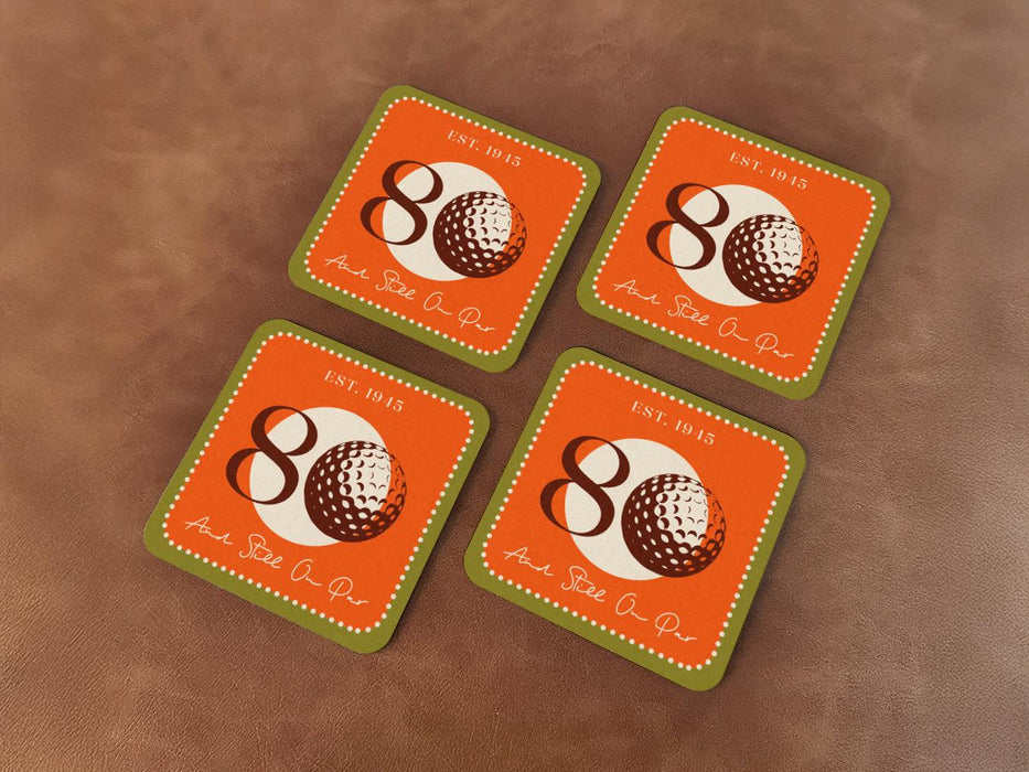 Custom square pulpboard coasters are shown sitting on leather surface. The coaster features design with illustrated golf ball, custom age and year, and vivid orange, cream, and green background.