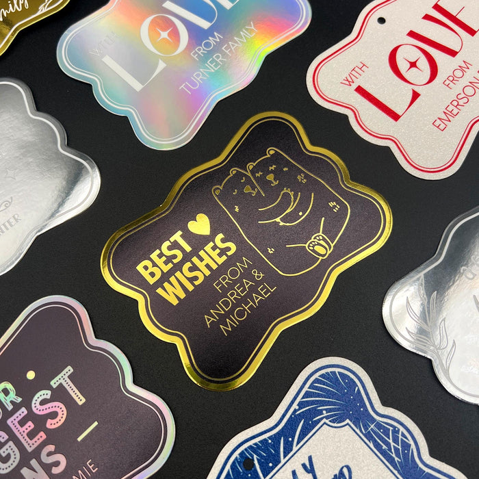 Assorted personalized wedding gift tags displayed on a dark background, featuring custom foil designs in multiple styles and colors including gold, rainbow, silver, and glitter finishes. The tags showcase elegant wedding-themed messages and customizable sender names.