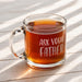 Clear glass mug filled with tea on a light wooden table with sunlight streaming across the surface. The warm amber tea adds a cozy and relaxed gift presentation.