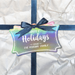 A slim rectangle gift tag with a blue ribbon, tied in a bow, attached to crumpled white paper. The tag has a UV printed message that reads "Happy Holidays".