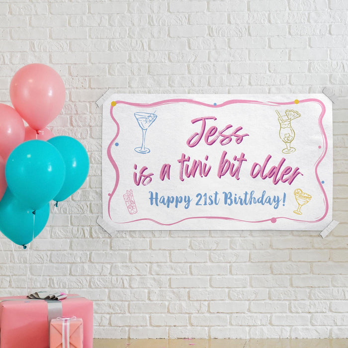 Personalized "Tini Bit Older" banner displayed on a white brick wall with colorful balloons and party decorations.