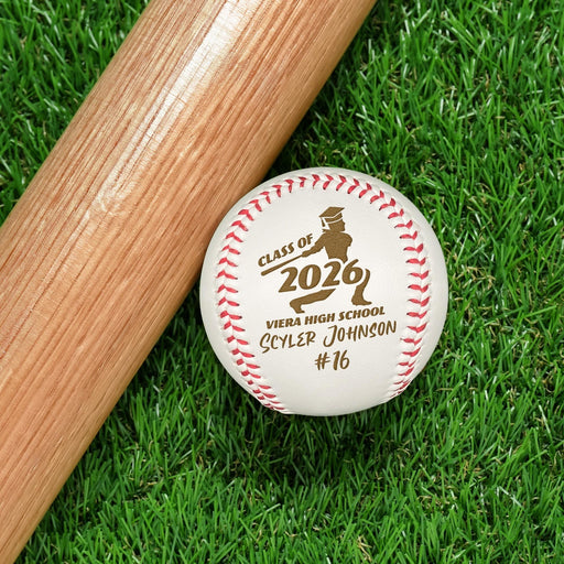 A baseball bat and a personalized baseball with text and a graphic on it are placed on a grassy surface. The design features a class year, a school name, a name and a jersey.