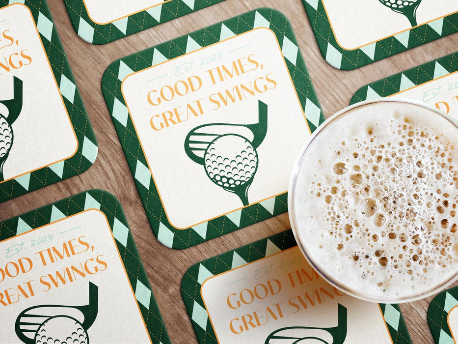 Custom square pulpboard coasters are shown laid out on a wooden surface. A beer mug is shown on top of a few of the coasters. The coaster features design with an illustration of a golf ball and with club behind it, argyle background pattern, and custom text.