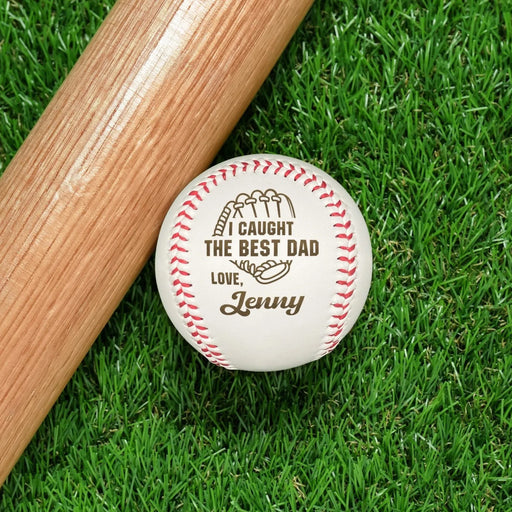 Baseball sits on grass next to baseball bat. Baseball has I Caught the Best Dad design engraved on it.