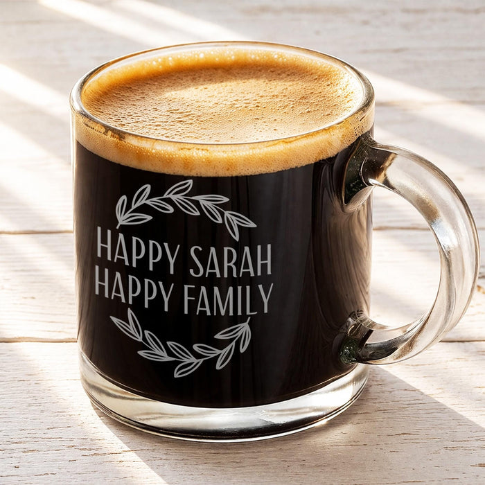 A glass mug filled with a dark liquid, likely coffee, with a wreath design engraved on its side.