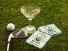 Custom square pulpboard coasters are shown sitting on a grassy, green golf course next to a martini glass, white golf ball, and golf club. The coaster features design with illustrated golf ball, club, and flag on top with wreath surrounding it, argyle background pattern, and custom text.