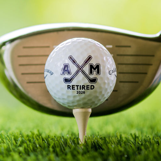 Close-up of a personalized retirement golf ball featuring crossed golf clubs, custom initials, and retirement year. Displayed on a tee in front of a golf club.