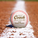 A personalized baseball rests on a dirt field, with a white line visible in the background.