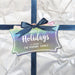 Slim rectangle personalized Happy Holidays hang tag attached to a white wrapped Christmas gift with a navy blue ribbon and bow.