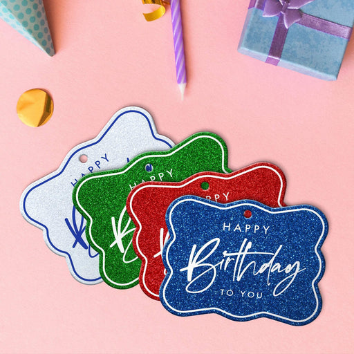 Four elegant script birthday hang tags in glitter blue, glitter red, glitter green, and glitter white lying beside colorful gifts and party decorations.