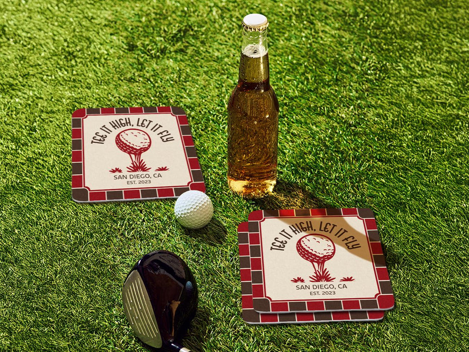 Custom square pulpboard coasters are shown sitting on a grassy, green golf course next to a bottle of beer, white golf ball, and golf club. The coasters feature design with an illustration of a golf ball on a tee, checkerboard border, and custom text.