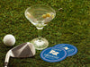 Custom round pulpboard coasters are shown sitting on a grass with a martini glass, golf club, and golf ball around them. The custom coasters feature design printed with blue background, pink circular border, and a beige illustrated golf flag.