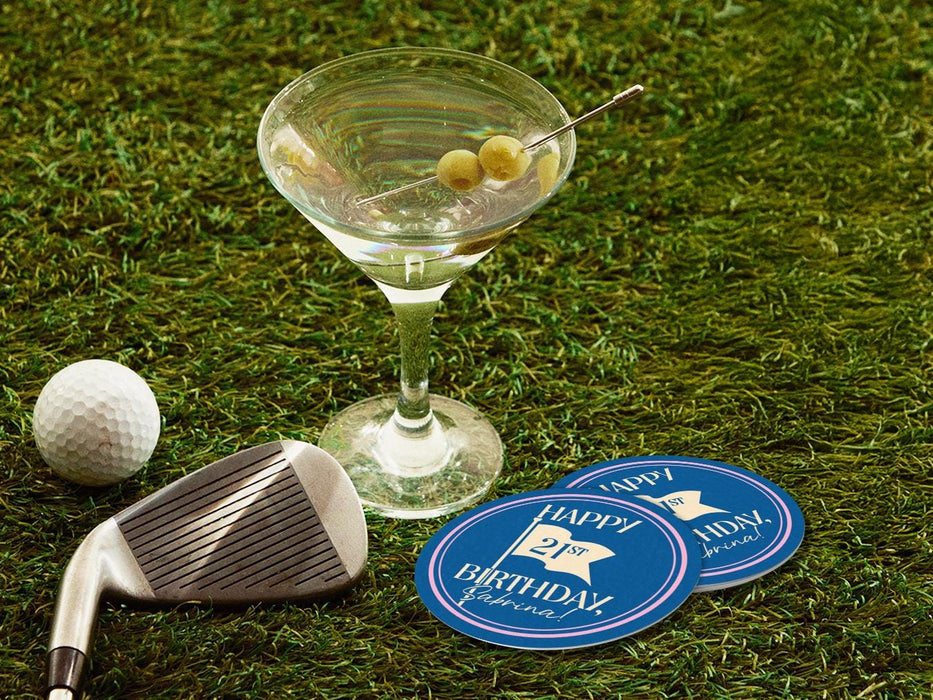 Custom round pulpboard coasters are shown sitting on a grass with a martini glass, golf club, and golf ball around them. The custom coasters feature design printed with blue background, pink circular border, and a beige illustrated golf flag.