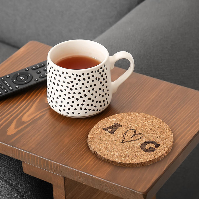 Personalized cork coaster with hand-drawn heart initials beside a coffee mug on a wood table.