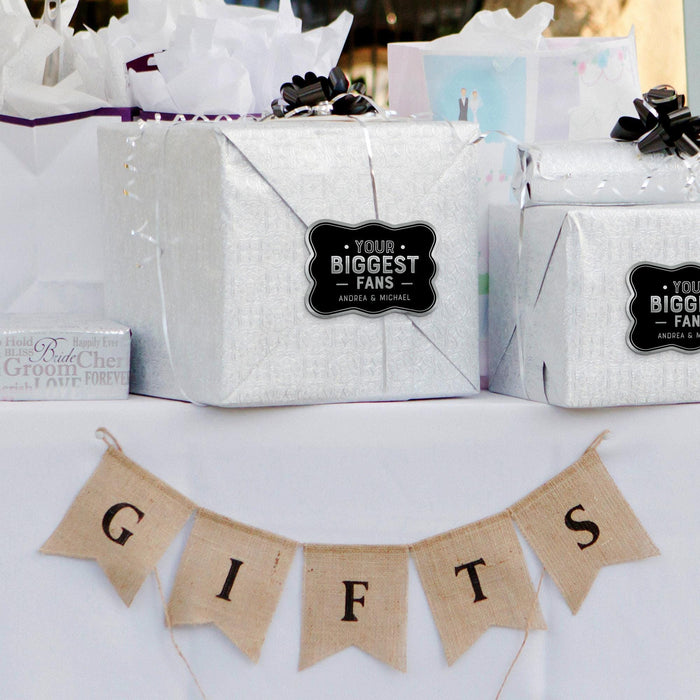 Silver gift bag decorated with a personalized “Your Biggest Fans” wedding gift tag in metallic silver foil. Styled with wedding décor and hanging banner accents for a romantic presentation.