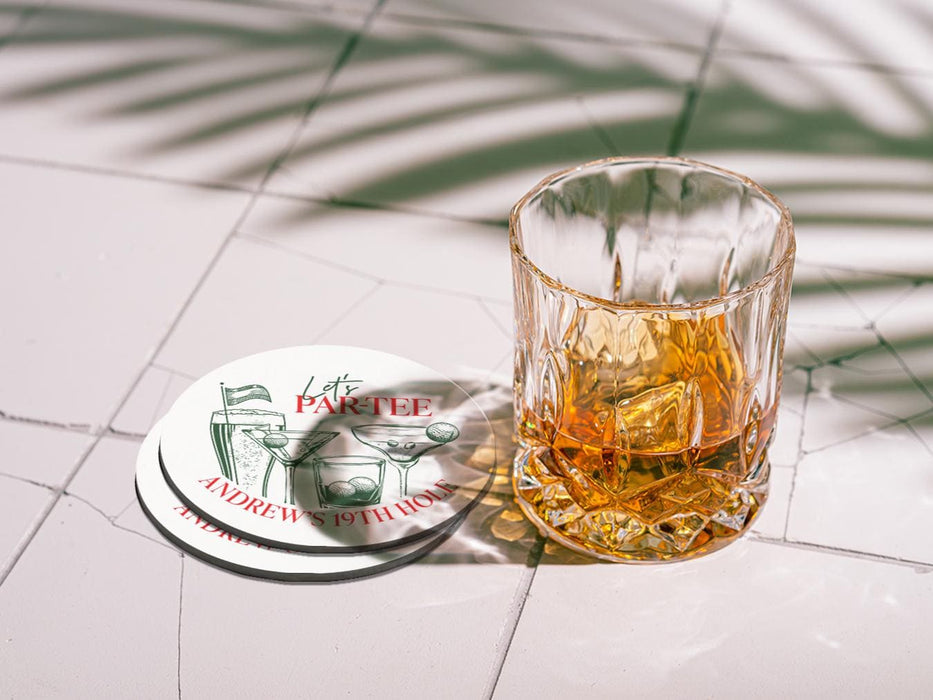 Custom square pulpboard coaster are shown laid out on marble floor. A whiskey glass is shown next to the coaster. The custom coasters feature design printed with green sketched cocktails with golf balls and flags within the drinks, and red and green text.
