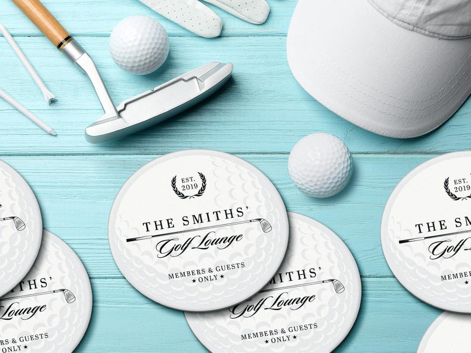 Custom round pulpboard coasters are shown sitting on a bright blue wooden surface. Golf balls, white golf tees, a golf club, and golf hat are shown next to the coasters. The custom coasters feature a golf club and overlayed golf ball design with the text Members & Guests Only between stars. The last name, club name, and established year can be customized.