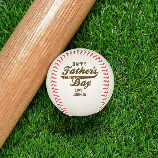 Baseball and wooden bat sits on grass. Baseball has Happy Father's Day design engraved on it.