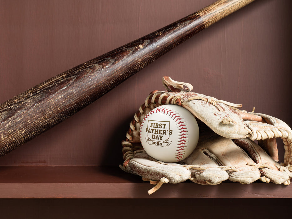 Baseball sits in glove on a shelf below a baseball bat. Baseball has First Father's Day design engraved on it.