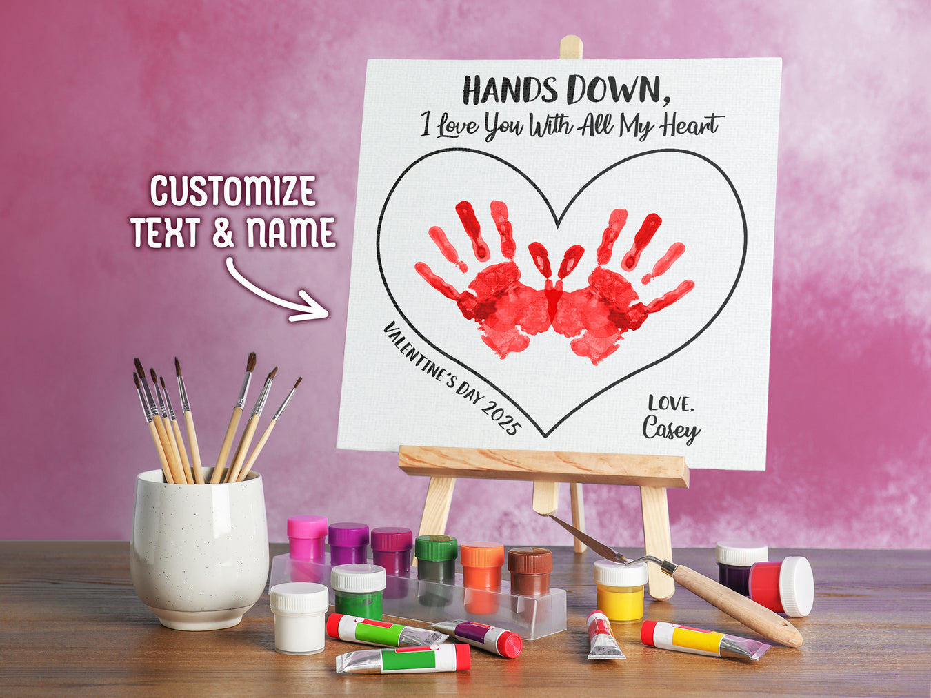 canvas panel with a custom Valentines Day design printed on it sitting on wooden easel, placed on a wooden table surrounded by painting supplies such as paint brushes, tubes and paint palettes, palette knife, in front of a pink wall