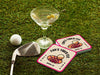 Custom square pulpboard coasters are shown sitting on a grassy, green golf course next to a martini glass, white golf ball, and golf club. The coaster features design with illustrated golf ball and hole, custom name, age, year, and location, and vivid orange, cream, and pink color palette.