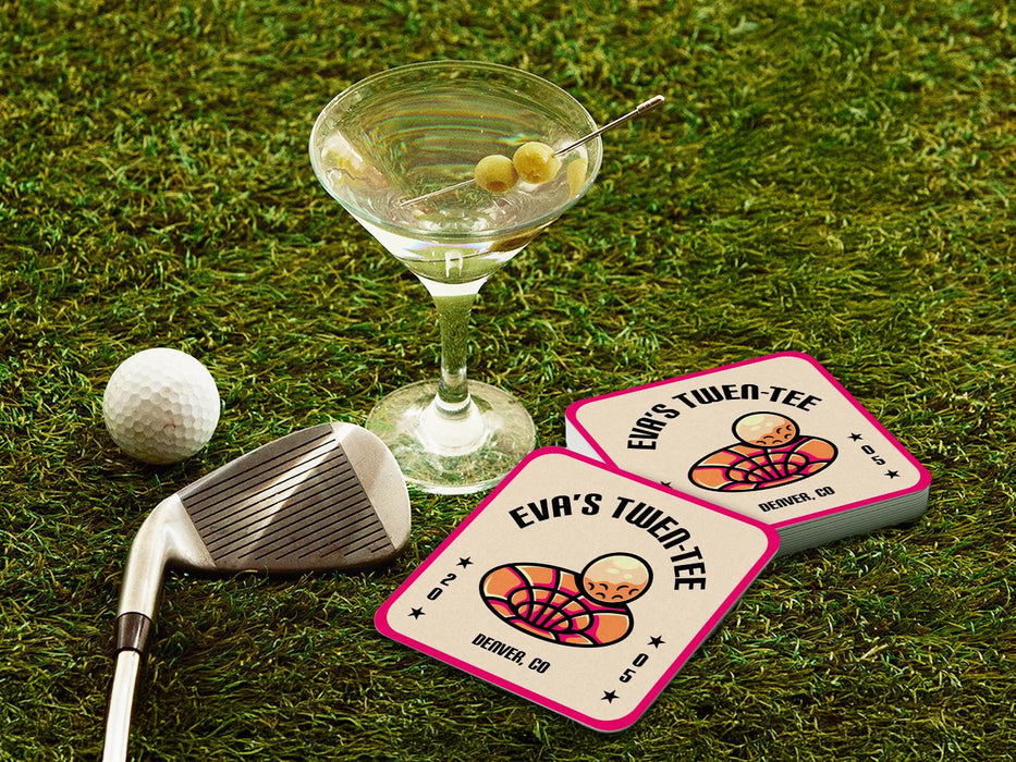 Custom square pulpboard coasters are shown sitting on a grassy, green golf course next to a martini glass, white golf ball, and golf club. The coaster features design with illustrated golf ball and hole, custom name, age, year, and location, and vivid orange, cream, and pink color palette.