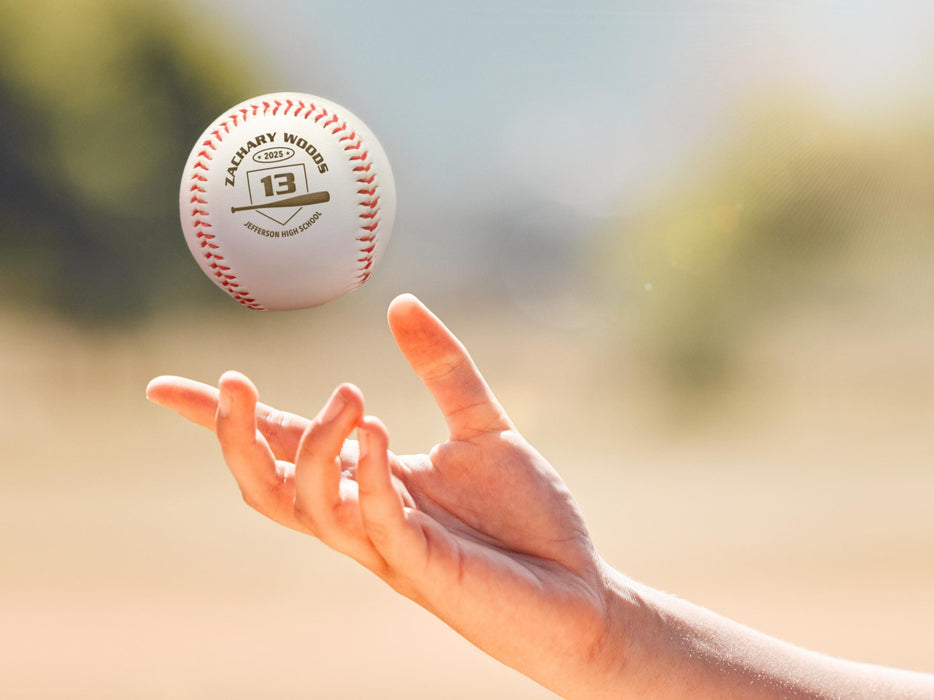 A person tosses a baseball into the air outside on a baseball field. The baseball has a year, player's name, number, and school name engraved into it. This design features a home plate and bat graphic with the text curved around it.