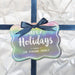 Rounded rectangle personalized Christmas hang tag attached to a white wrapped gift with a navy ribbon and bow.