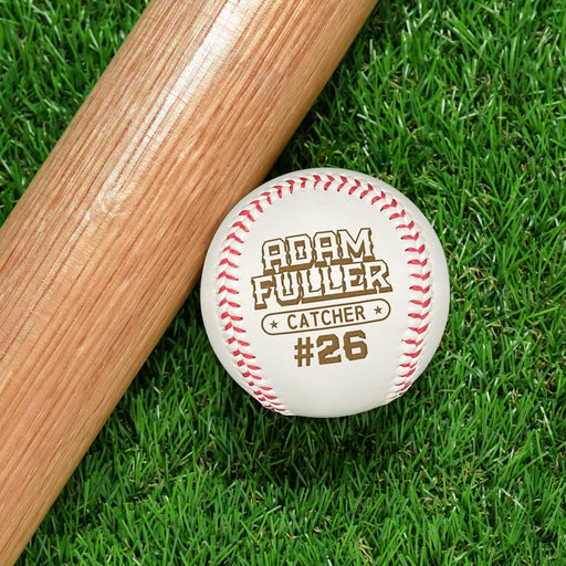 A personalized baseball with the name "Adam Fuller" engraved on it, along with the position "CATCHER" and the number "#26" printed below, rests on a grassy surface next to a wooden baseball bat.