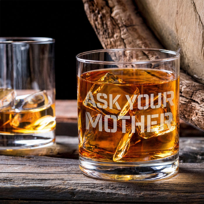 Lifestyle image of the whiskey glass with large ice cubes on a wooden table beside another drink. The “Ask Your Mother” design adds a fun and casual bar feel.
