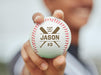 A baseball player holds up an engraved baseball outside. This engraved design features a bold crossed bat graphic with a baseball player's graduating year, first name, and jersey number.