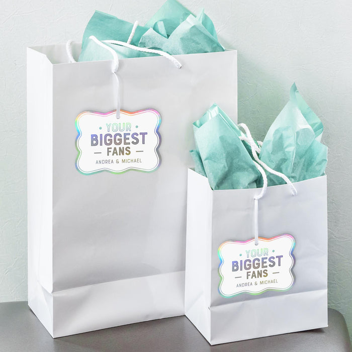 White gift bags with tissue paper featuring personalized “Your Biggest Fans” wedding gift tags in holographic rainbow foil. A fun and modern packaging idea for wedding and bridal shower gifts.