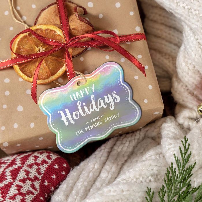 Rounded rectangle personalized Christmas hang tag tied to a kraft paper gift with red ribbon, dried orange slices, greenery, and a cozy knit background.