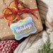 Rounded rectangle personalized Christmas hang tag tied to a kraft paper gift with red ribbon, dried orange slices, greenery, and a cozy knit background.