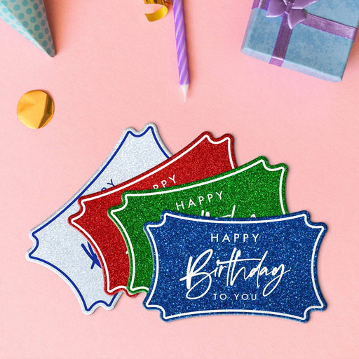 Four birthday gift tags in glitter red, glitter blue, glitter green, and glitter white lying beside colorful gifts and party decorations.