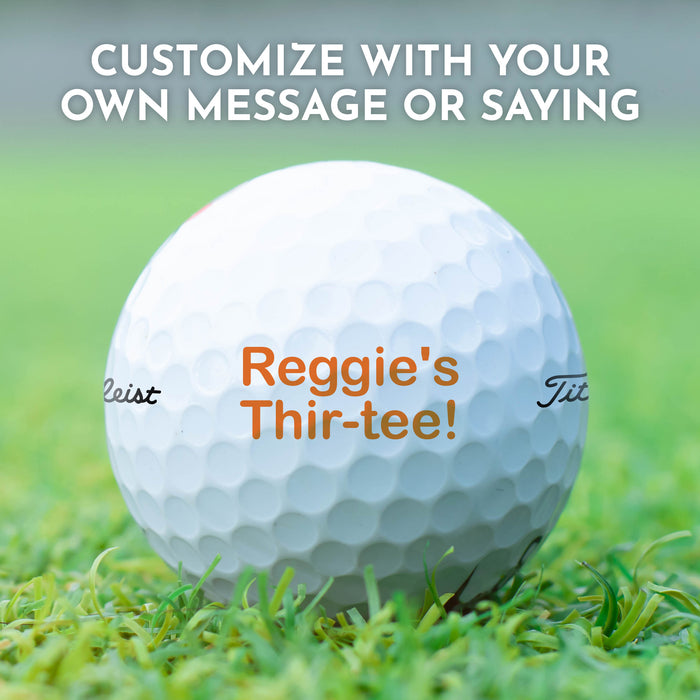 A golf ball with the text "Reggie's Thir-tee!" printed on it, placed on a grassy field.