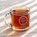 A glass mug filled with a warm beverage, likely tea, sits on a wooden surface. The mug has a personalized message engraved on it, which reads "Happy Father's Day Love, Daniel".