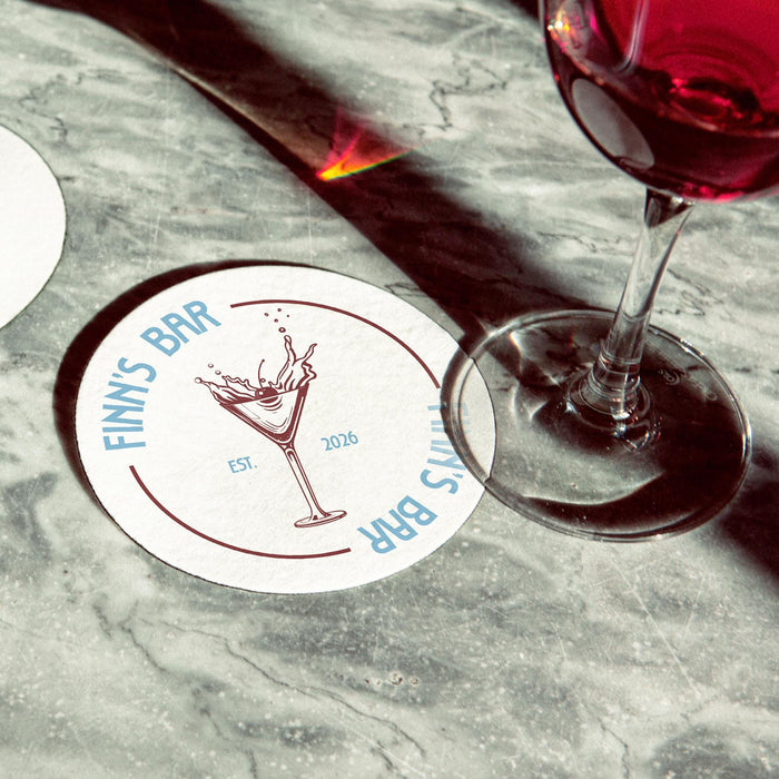 Personalized martini coaster placed beside cocktail glasses on a marble bar surface, featuring the custom bar name, martini illustration, and established year.