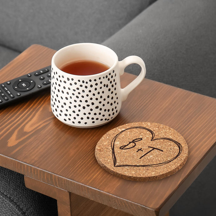 Personalized cork coaster with a large hand-drawn heart initial design beside a coffee mug on a wood table.