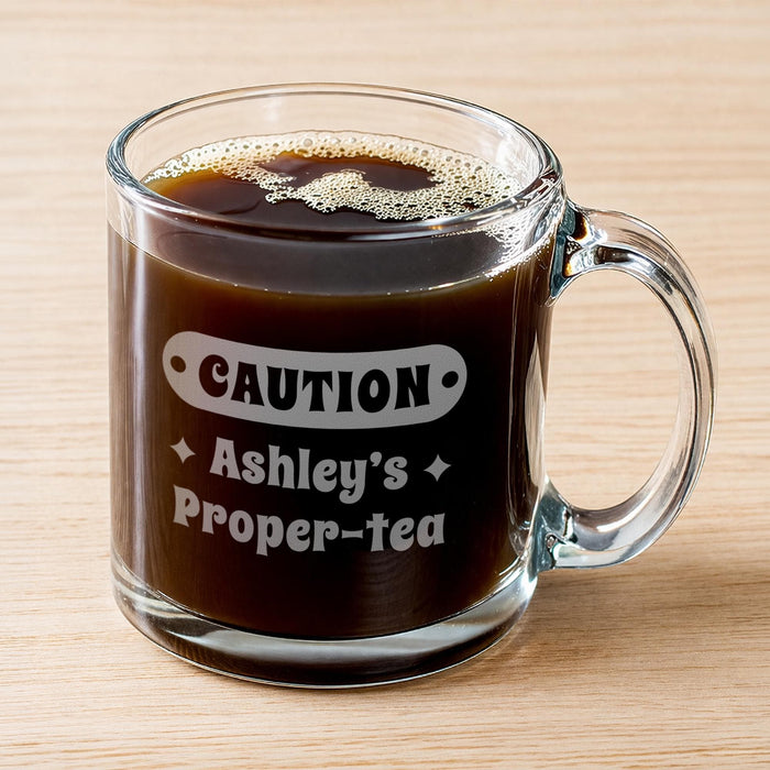 A glass mug filled with a liquid. The mug has a handle and an engraving that says "CAUTION Ashley's Proper-Tea".