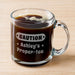 A glass mug filled with a liquid. The mug has a handle and an engraving that says "CAUTION Ashley's Proper-Tea".