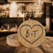 Personalized cork coasters with a large hand-drawn heart monogram and initials displayed in a rustic bar setting.