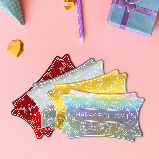 Four floral birthday gift tags in metallic gold, metallic silver, metallic red, and metallic rainbow lying beside colorful gifts and party decorations.