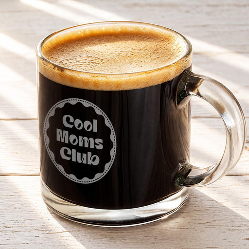 A glass mug filled with a dark liquid, likely coffee, with a foamy top, sitting on a wooden surface.