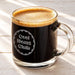 A glass mug filled with a dark liquid, likely coffee, with a foamy top, sitting on a wooden surface.